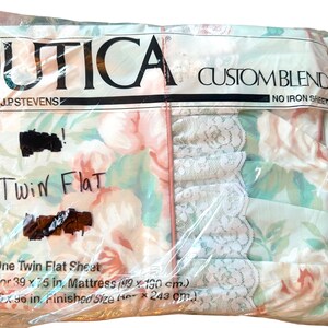 Könnte beinhalten: Ein flaches Bettlaken für Einzelbetten von Utica J.P. Stevens mit Blumenmuster, rosa Rosen und grünen Blättern auf hellem Grund. Die Verpackung zeigt weiße Spitzenborte, den Text "UTICA CUSTOM BLEND NO IRON SHEET" und Maße für eine 99 x 190 cm Matratze.