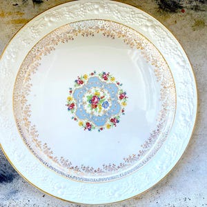 May include: A white ceramic serving bowl with a raised rim and gold trim. The bowl features a floral design with blue, pink, yellow, and green flowers.