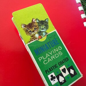 May include: A green and white box of "MINIATURE PLAYING CARDS" with a picture of two kittens on the top card. The box is on a red surface. The text "PLASTIC COATED" is printed on the box.
