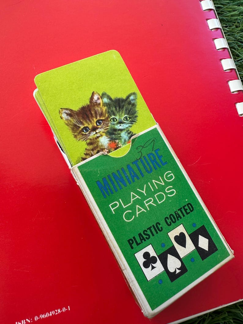 May include: A green and white box of "MINIATURE PLAYING CARDS" with a picture of two kittens on the top card. The box is on a red surface. The text "PLASTIC COATED" is printed on the box.