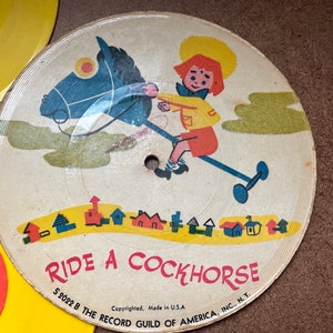 May include: A vintage children's record with a yellow label and a colorful illustration of a child riding a rocking horse. The text "RIDE A COCKHORSE" is printed in red on the record. The record is labeled "S 2022 B THE RECORD GUILD OF AMERICA, INC., N.Y."