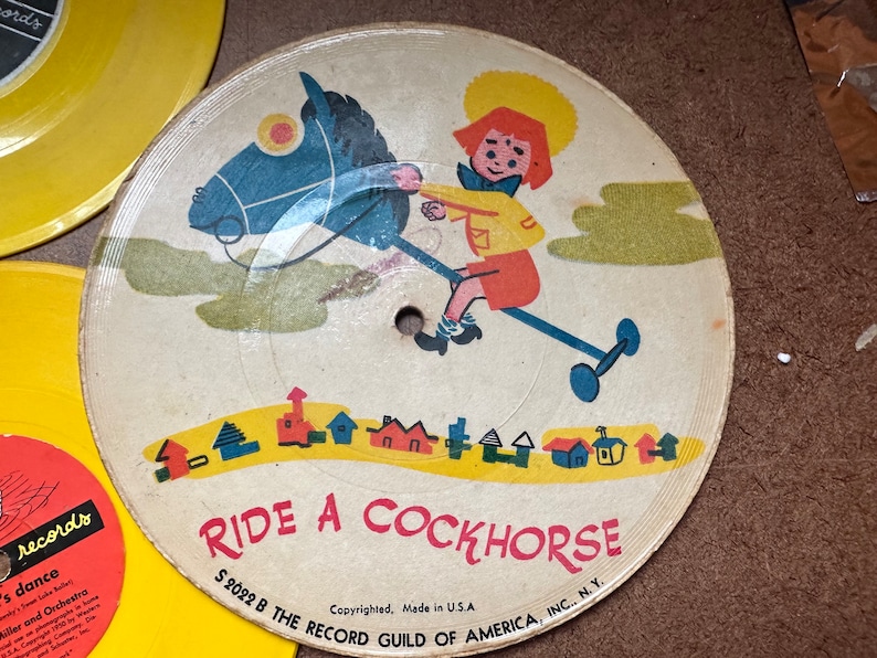 May include: A vintage children's record with a yellow label and a colorful illustration of a child riding a rocking horse. The text "RIDE A COCKHORSE" is printed in red on the record. The record is labeled "S 2022 B THE RECORD GUILD OF AMERICA, INC., N.Y."