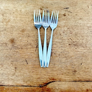 May include: Three silver forks with slender handles and four tines each are arranged on a wooden surface. The forks have a simple, elegant design, suitable for everyday dining. The wood has a warm, natural tone.
