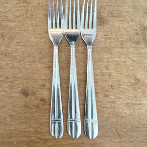 May include: Three silver forks with tines pointing upwards are arranged vertically on a wooden surface. The forks have a simple, elegant design with a rounded base and a slightly tapered handle. The wood grain is visible in the background.