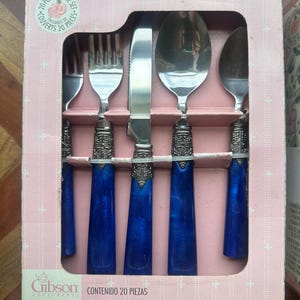 May include: A 20-piece flatware set in a pink box featuring a fork, knife, and spoons with metallic silver heads, ornate silver-toned collars, and deep blue marbled handles. The packaging includes the text 20 PC. FLATWARE SET and Gibson.