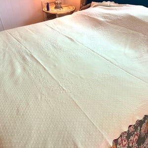 May include: Ivory-colored bedspread with a scalloped edge. The bedspread features a textured pattern of small dots and floral embroidery along the borders. The bedspread is spread over a bed with a floral patterned bed skirt.