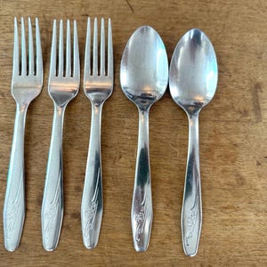 May include: Five pieces of silverware, including three forks and two spoons, are arranged on a wooden surface. The silverware is silver-colored with a floral design on the handles. The forks have four tines.