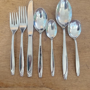 May include: A set of polished silver flatware, including forks, knives, and spoons of varying sizes. The silverware is arranged on a wooden surface, reflecting the light. The handles have a decorative, curved design.