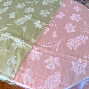 May include: Two tablecloths, one pale green and one pink, are displayed. Both feature a repeating pattern of white flowers, butterflies, and stems. The fabric has a subtle sheen, suggesting a satin or similar material. The floral design adds a touch of elegance.