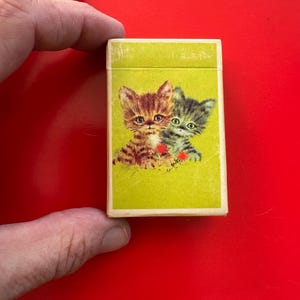 May include: A vintage matchbox with a lime green background featuring two kittens. One kitten is brown and the other is gray. The matchbox has the ISBN 0-9604928-0-1 printed on the bottom.