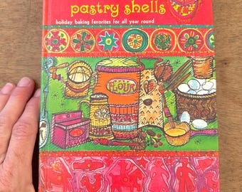 May include: A red, green, and yellow cookbook titled "Jingle Bells and Pastry Shells" by Alma Smith Payne. The cover features illustrations of holiday baking ingredients and a festive design.