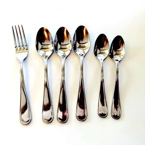 May include: A set of stainless steel silverware, including a fork and five spoons of varying sizes. The utensils are arranged on a white surface, reflecting light and creating a bright, clean aesthetic. Ideal for dining.