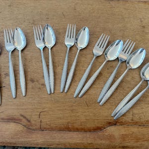 May include: A collection of stainless steel silverware, including spoons and forks, arranged on a wooden surface. The silverware has a simple, elegant design with a textured pattern on the handles. The spoons and forks are of standard size.