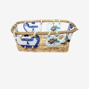 May include: A rectangular woven wicker basket featuring a wooden rim. Attached to the sides are four ceramic pieces: two white teapots with blue patterns and two white pitchers decorated with colorful floral motifs, all secured with twine.