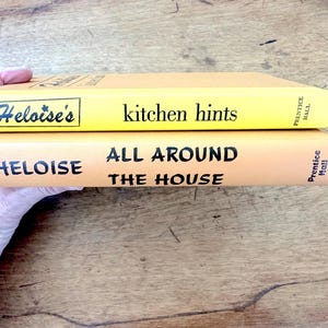 May include: Two yellow paperback books stacked on top of each other. The top book has the title "Heloise's kitchen hints" and the bottom book has the title "Heloise All Around The House". Both books have the publisher "Prentice Hall" printed on the spine.