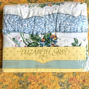 May include: A twin flat sheet by Elizabeth Gray Color Forum in plastic packaging. The white sheet features a floral pattern with blue, green, and yellow tones, accented by a white eyelet lace trim and a blue and white leaf-patterned ruffle.