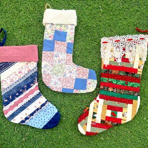 May include: Three handmade Christmas stockings on green grass. The stockings feature patchwork designs in various colors, including blue, red, and white. One stocking has a pink cuff, another has a white fluffy cuff, and the third has a lace trim.
