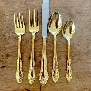 May include: A set of gold-plated silverware, including a knife, fork, and two spoons. The silverware has an ornate design and is laid out on a wooden surface.