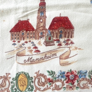 May include: Cream-colored textile featuring a vintage illustration of Mannheim, Germany. The design includes buildings with red roofs, a tall clock tower, and a banner reading "Mannheim." A floral border with red, blue, and green accents completes the design.
