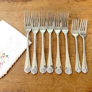 Op de afbeelding: Acht zilveren vorken met een schelpontwerp op de handvatten, gerangschikt op een houten oppervlak. Een wit geborduurd zakdoek met een bloemenmandontwerp staat op de achtergrond.
