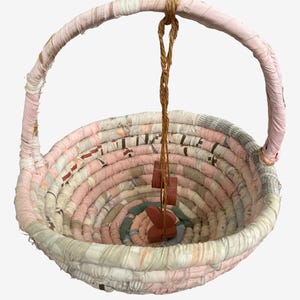 May include: A round basket constructed from coiled fabric strips in shades of pale pink, white, and muted grey. The basket features a single arched handle wrapped in matching fabric and a brown twine cord hanging from the center with small wooden shapes.