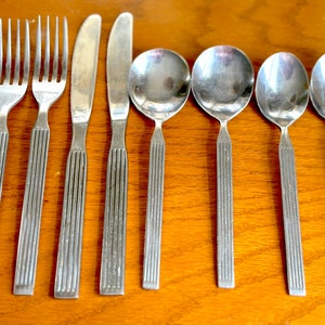 Peut inclure: Un ensemble de huit couverts argentés, comprenant trois fourchettes, un couteau et quatre cuillères. Les couverts ont un design strié et sont disposés sur une surface en bois.
