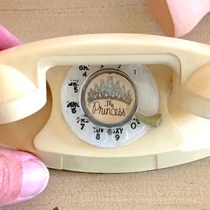 Könnte beinhalten: Ein weißes Vintage-Drehwahltelefon mit einem cremefarbenen Zifferblatt. Das Zifferblatt hat eine Krone und den Text "The Princess" darauf.
