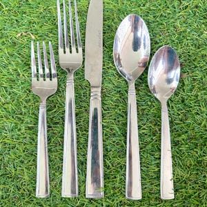 Könnte beinhalten: Ein Set aus fünf Edelstahl-Besteckteilen, darunter zwei Gabeln, ein Messer und zwei Löffel. Auf der Messerklinge steht "CROFTON". Das Besteck liegt auf einer grünen Grasfläche.