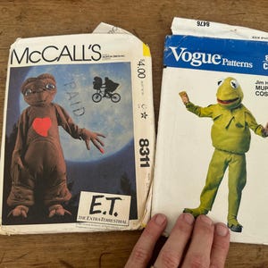 May include: Two vintage sewing pattern envelopes. The left envelope is McCall's 8311 featuring an E.T. the Extra-Terrestrial costume with a brown character suit and red heart. The right envelope is Vogue Patterns 8476 for a Kermit the Frog costume.