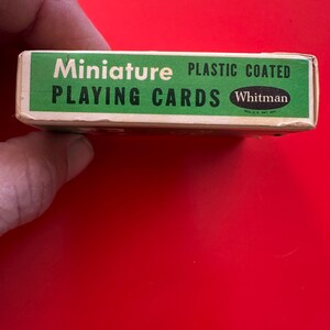 May include: A miniature deck of playing cards in a green and white box. The box reads "Miniature PLAYING CARDS" and "PLASTIC COATED" with the "Whitman" logo. The box is held by a hand against a red background.