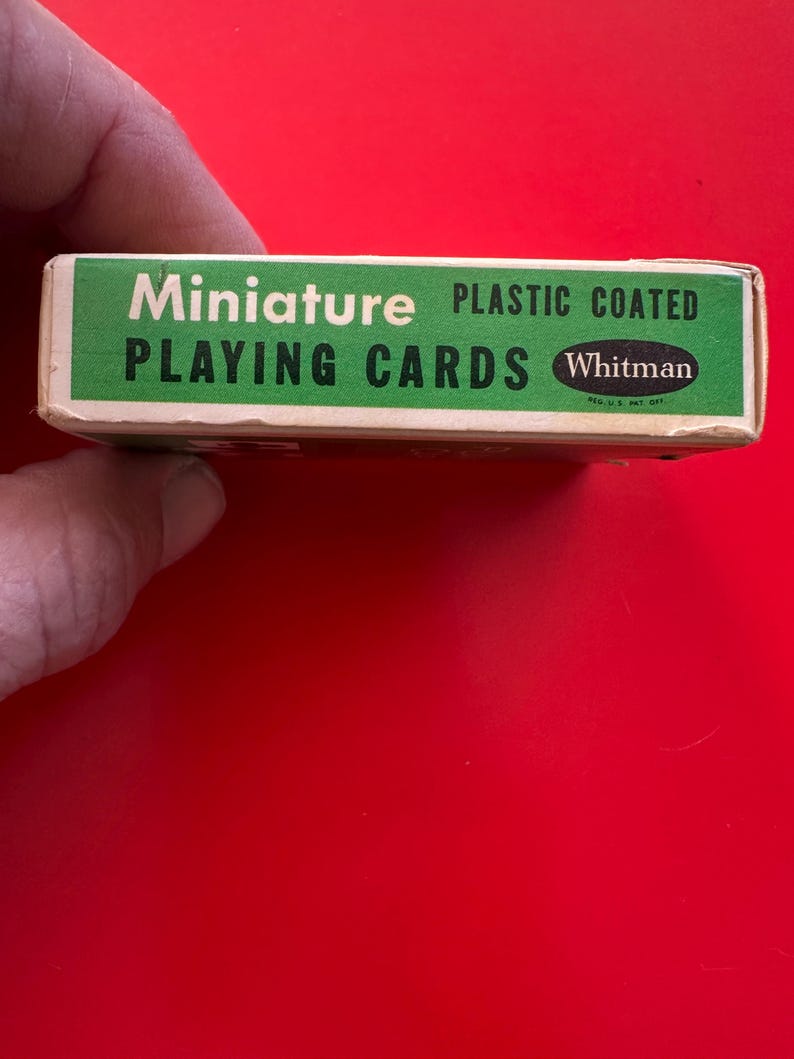 May include: A miniature deck of playing cards in a green and white box. The box reads "Miniature PLAYING CARDS" and "PLASTIC COATED" with the "Whitman" logo. The box is held by a hand against a red background.