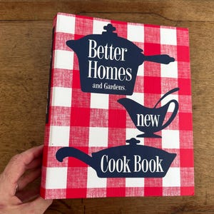 May include: A vintage cookbook titled "Better Homes and Gardens New Cook Book." The cover features a red and white checkered pattern with navy blue illustrations of cookware and text. The book is held by a hand.