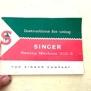 May include: A rectangular instruction manual for a Singer sewing machine model 201-2. The cover features a green top section, a red middle section, and a white bottom section with the text Instructions for using SINGER Sewing Machine 201-2.