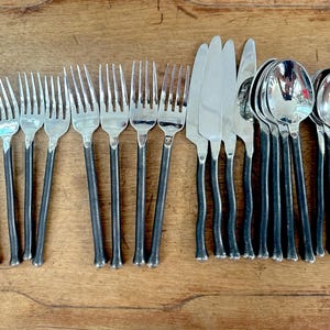 May include: A collection of stainless steel silverware, including forks, knives, and spoons, with dark gray handles. The utensils are arranged on a wooden surface, showcasing a complete dining set.
