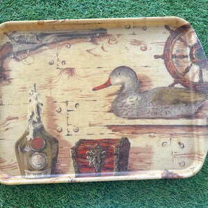 Può includere: Vassoio rettangolare con un design vintage. Il vassoio presenta un'anatra, un timone, una bottiglia, un forziere e una pistola su uno sfondo a venature di legno. Il vassoio ha angoli arrotondati e una finitura lucida.