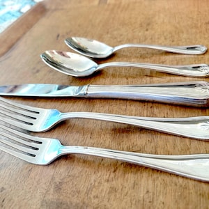 May include: A set of silver-colored flatware, including two forks, a knife, and two spoons, arranged on a wooden surface. The silverware has decorative detailing on the handles. The spoons are small, and the knife has a rounded tip.