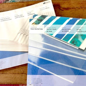 Könnte beinhalten: Zwei Packungen Scrapbook-Zubehör. Eine Packung enthält weiße Scrapbook-Seiten, die andere transparente Seitenschutzhüllen mit blauem und türkisfarbenem Design. Die Packungen liegen auf einer Holzoberfläche.