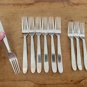 May include: A collection of stainless steel forks with a simple, elegant design. The forks have a long, slender handle and four tines. One fork is held by a hand. The forks are arranged on a wooden surface.