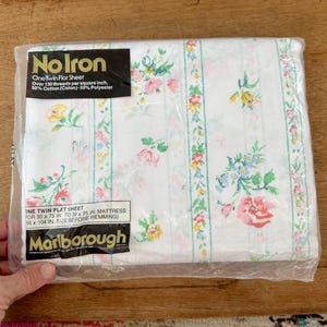 Könnte beinhalten: Ein verpacktes Twin-Flachbettlaken mit floralem Muster. Das Laken ist weiß mit vertikalen Streifen aus rosa, gelben und blauen Blumen. Die Verpackung enthält den Text "No Iron" und "Marlborough". Das Laken ist für eine Matratze von 76 x 191 cm bis 99 x 191 cm konzipiert.