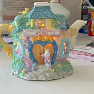 May include: A decorative, pastel-coloured teapot in the shape of a bunny hotel. It has a blue roof, a heart-shaped door, and a small bunny figurine. The word "HOTEL" is displayed on a sign above the door. The teapot is illuminated from within.