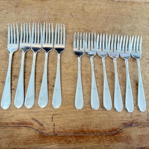 May include: A set of eleven silver-toned metal forks arranged in two rows. The five forks on the left are larger, while the six forks on the right are smaller. Each fork features a slender handle with a subtle, curved decorative line detail.