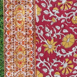 May include: Close-up of a textile with a floral design. The fabric features a red background with yellow flowers and white vines. A patterned border with orange and yellow floral motifs runs along the left side, creating a vibrant and detailed pattern.