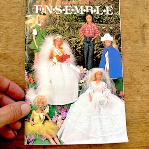 May include: A vintage "Fashion Doll Ensemble" magazine cover featuring several dolls in various outfits. The dolls are dressed in a variety of styles, including a wedding dress, a scout uniform, and a blue cape. The cover has a floral background.