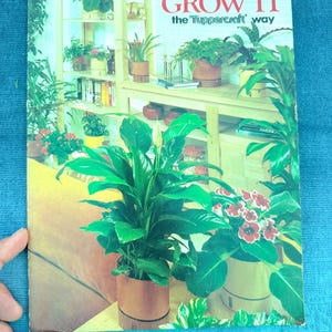Può includere: Una copertina di libro vintage con il titolo "You Can Grow It the Tupperware Way". La copertina presenta una foto di un soggiorno con diverse piante in vaso, tra cui un giglio della pace, una pianta a fiori rossi e una felce.