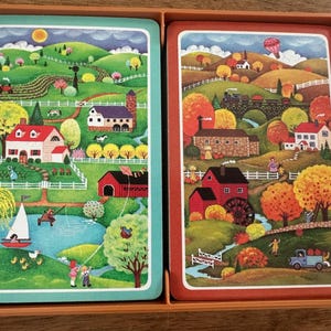 Puede incluir: Un juego de dos barajas de cartas ilustradas en una caja marrón. Una baraja presenta una escena de primavera con una casa roja, un velero y un estanque. La otra baraja representa una escena de otoño con un tren, un granero rojo y un camión.