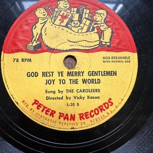 May include: A vintage 78 RPM record with a red, yellow, and black label. The label features a cartoon illustration of children carrying a bag of gifts. The record is titled "God Rest Ye Merry Gentlemen Joy To The World" and is sung by The Caroleers. The record is directed by Vicky Kasen and is a Peter Pan Records release.