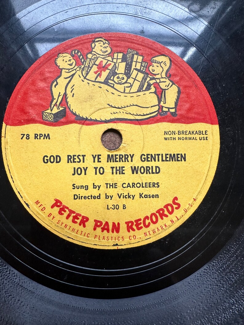 May include: A vintage 78 RPM record with a red, yellow, and black label. The label features a cartoon illustration of children carrying a bag of gifts. The record is titled "God Rest Ye Merry Gentlemen Joy To The World" and is sung by The Caroleers. The record is directed by Vicky Kasen and is a Peter Pan Records release.