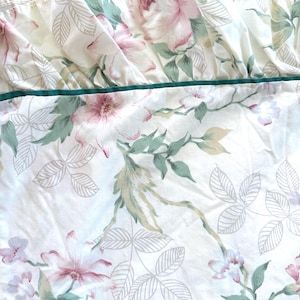 May include: White floral bed linens with a ruffled edge. The pattern features pink and purple flowers with green leaves and stems. A dark green trim accents the edge of the linens. The fabric is soft and appears to be cotton.