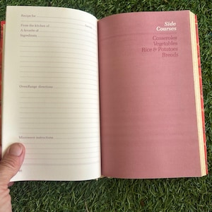 May include: Open cookbook with lined pages for recipes and a pink page with "Side Courses" and food categories. The book is held open, revealing the interior pages. The cover is red.