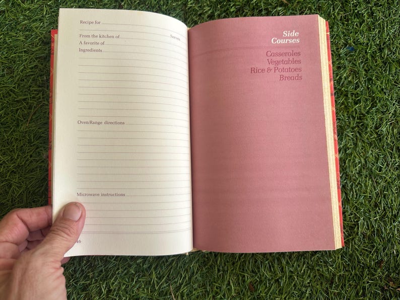 May include: Open cookbook with lined pages for recipes and a pink page with "Side Courses" and food categories. The book is held open, revealing the interior pages. The cover is red.
